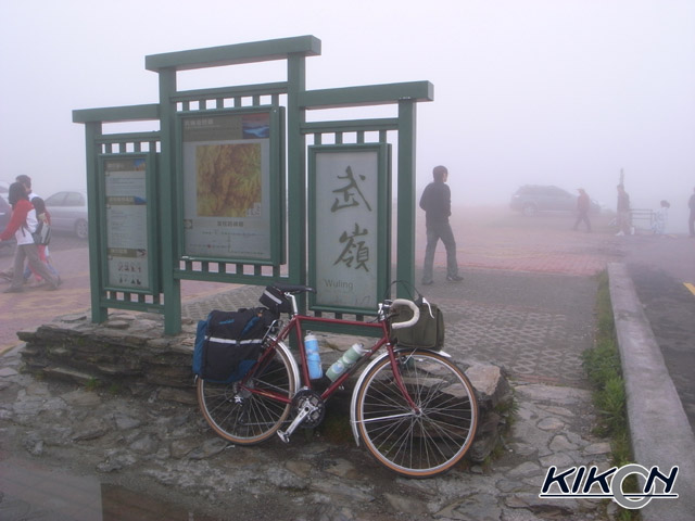 霧のなかに「武嶺」と書かれた案内板、その足下に自転車を停めている