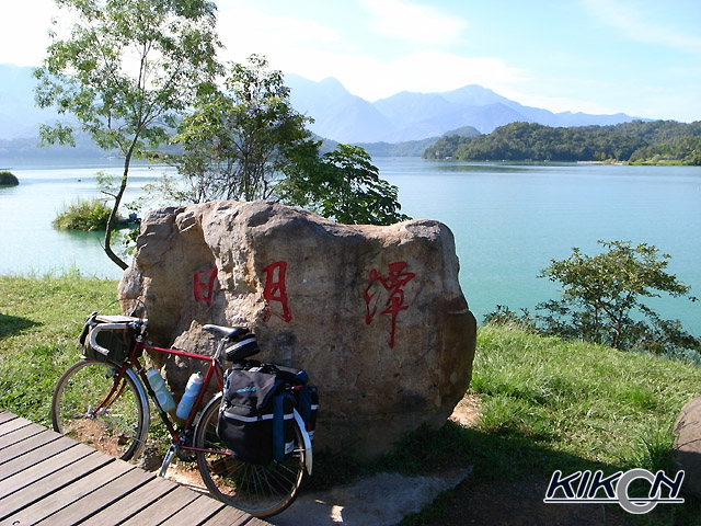 「日月潭」と彫られた岩の碑の前に自転車を停めている、背景には穏やかな大きな湖が広がっている。遠くに山々がそびえている