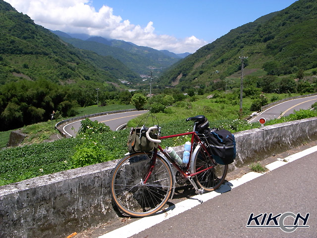 車道の脇に自転車を停めている。背後には山々が広がり、曲がりくねった山道が見える