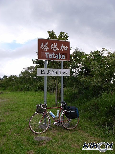 木々の密集した薮の脇に立つ、「塔塔加Tataka」「標高2610公尺」とかかれた表示版の前に自転車を停めている