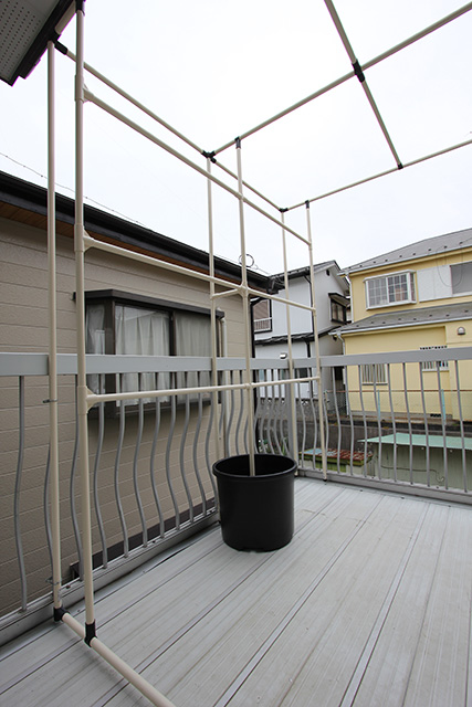 ベランダにある藤棚状パイプ組の一辺に支柱がたてられ、ヨコに3本のパイプが渡されている。支柱の1本は大きなポットの中から立ち上がっている