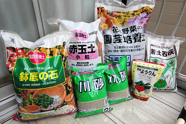 ベランダに、袋に入っている土石や砂などがおかれている。「鉢底の石」「川砂」「苦土石灰」「BMようりん」「赤玉土」がある。